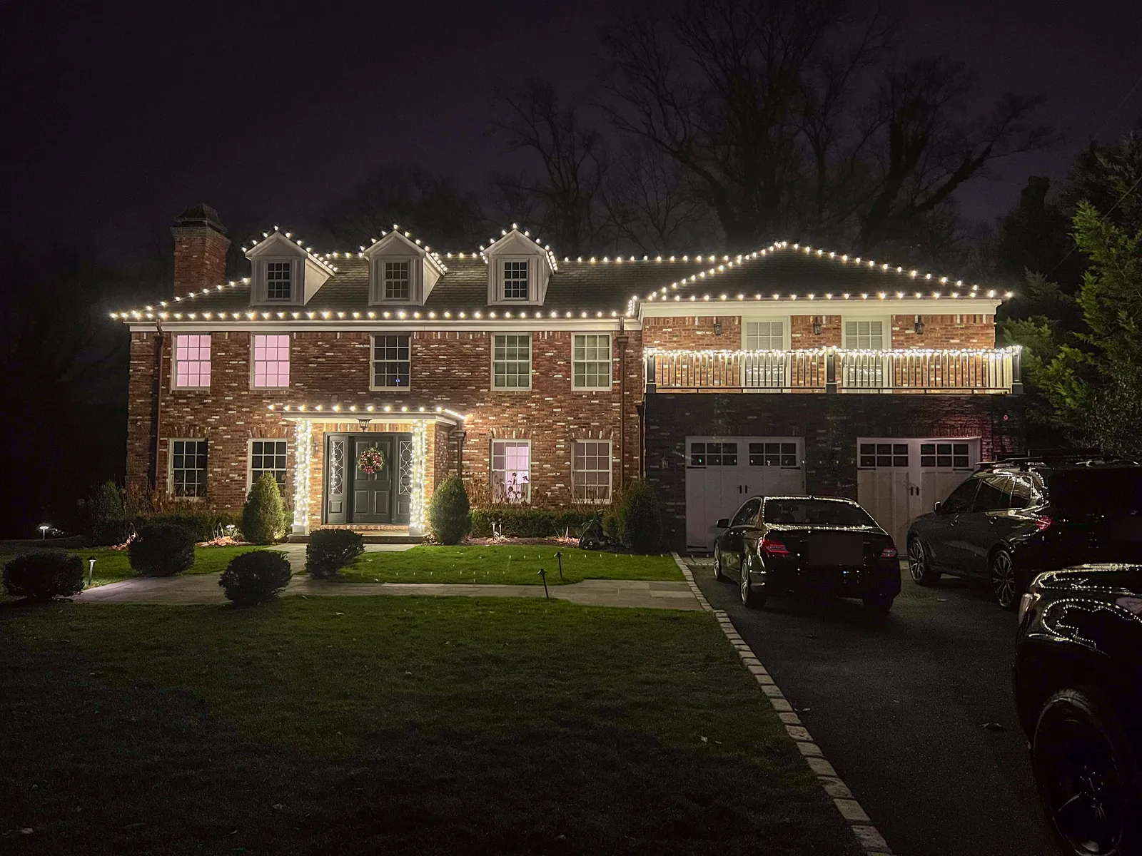 Brick colonial in Manhasset at night with roofline lights on three dormers, a lit balcony railing and a wreath on the door