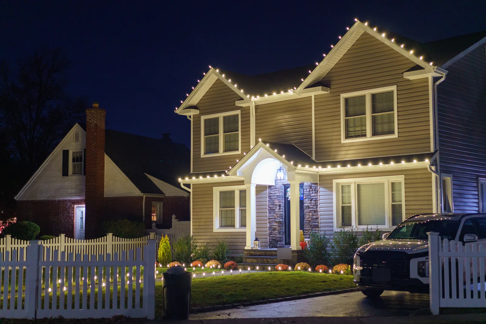 Two story colonial in Franklin Square at night with warm white roofline lights, a lit portico and stake lights along a picket fence