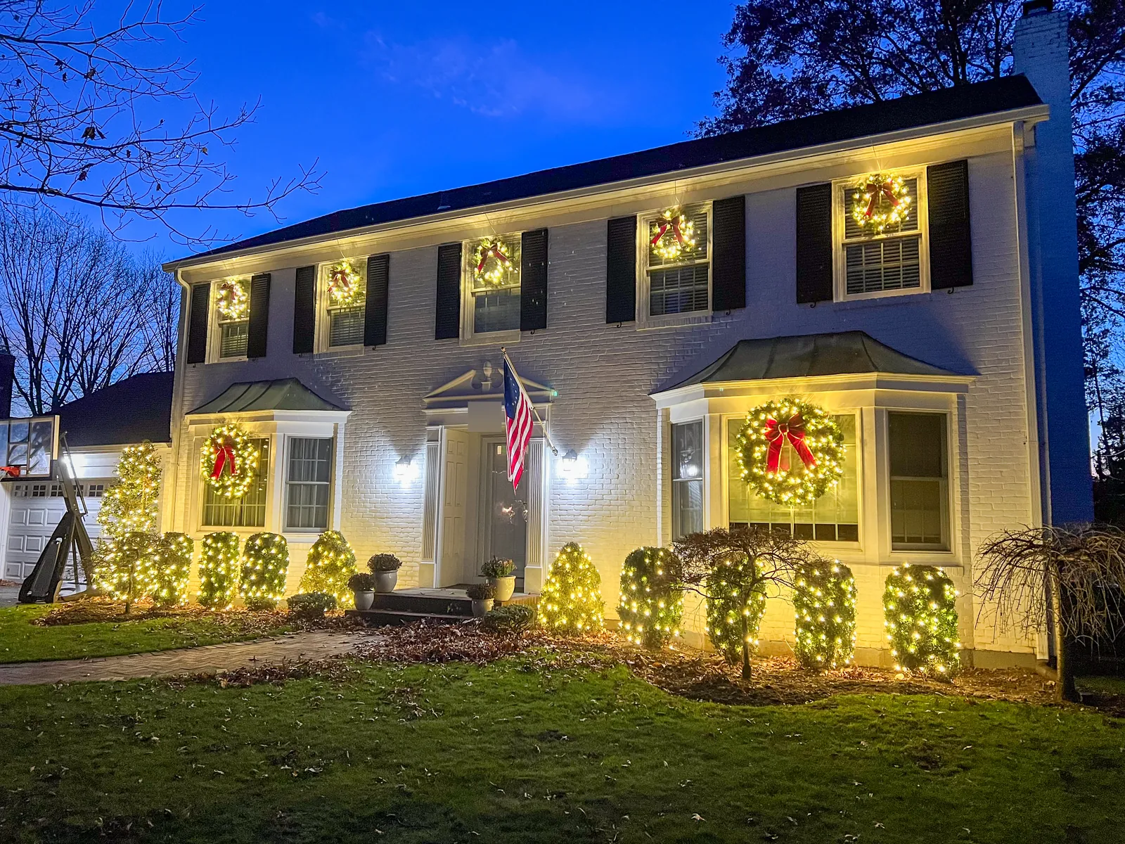 White brick colonial in Garden City at dusk with lit wreaths on every window and shrubs wrapped in warm white lights